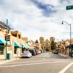 A street view of Old Santa Fe Trail, showing adobe buildings, a cathedral in the distance, and street signs under a blue sky.