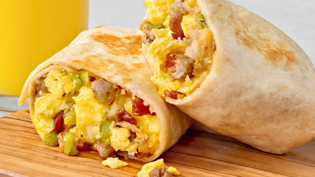 A breakfast burrito cut in half to show fillings of scrambled eggs, sausage, bacon, and green peppers on a wooden board.