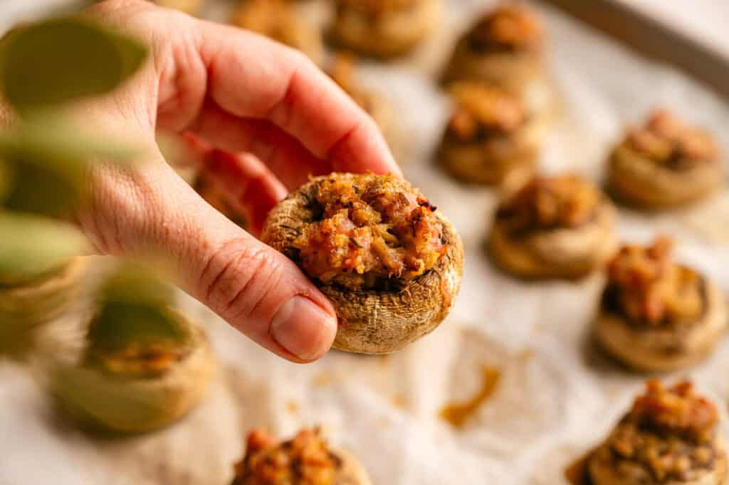 A hand holds a single stuffed mushroom, with several more on a parchment-lined baking sheet in the background.