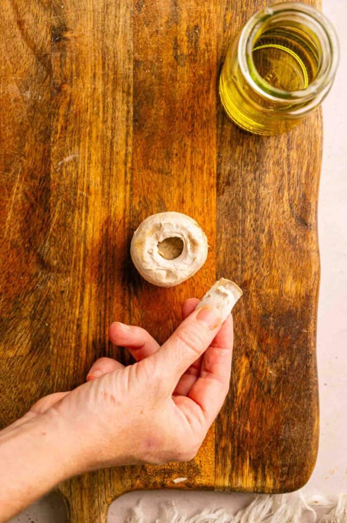 A hand places a mushroom stem onto a wooden cutting board next to a mushroom cap and a small jar of oil.