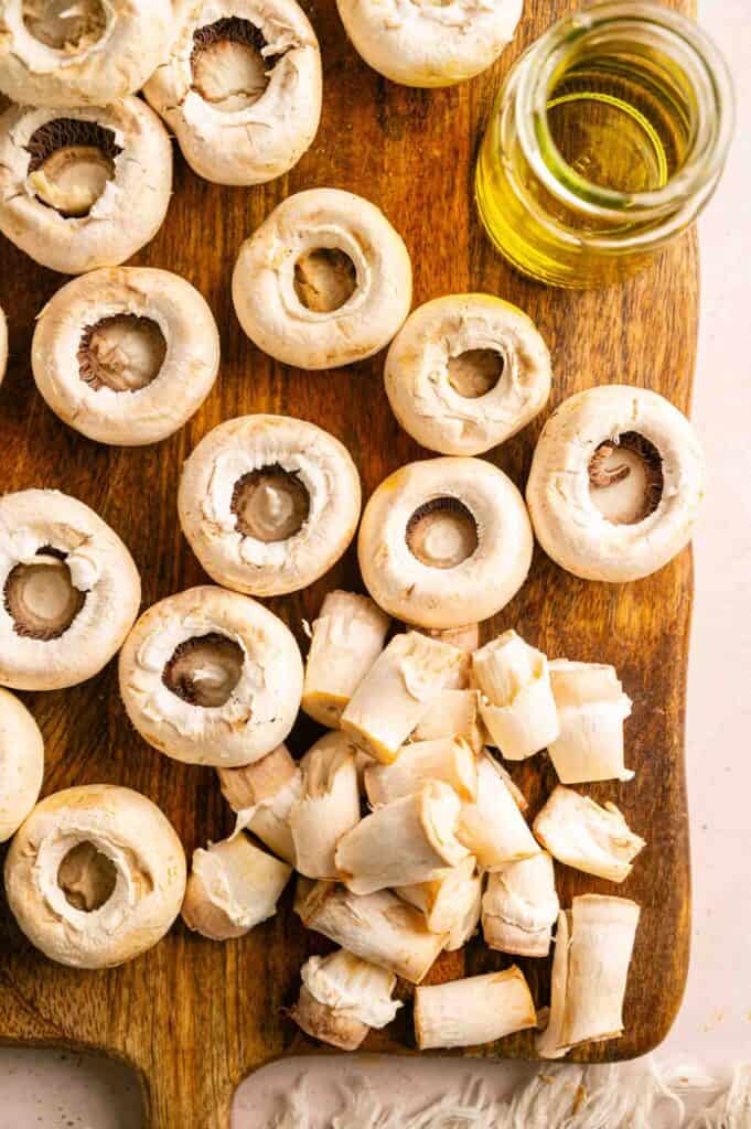 Whole white mushrooms and chopped mushroom stems on a wooden cutting board next to a small glass jar of olive oil.