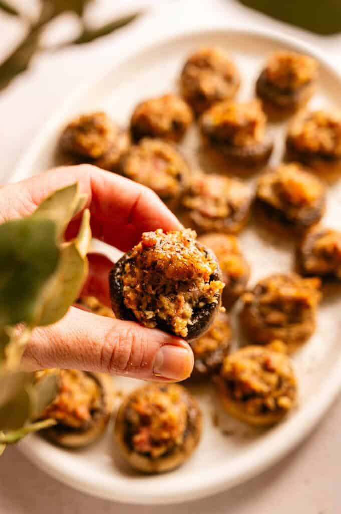 A hand holding a single stuffed mushroom, with a platter of additional stuffed mushrooms in the background.