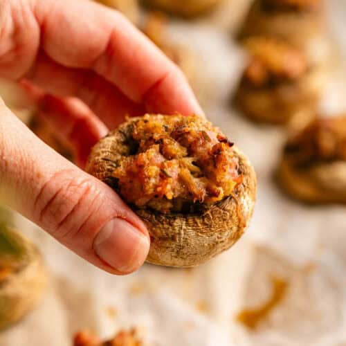 A hand holding a single stuffed mushroom, with more prepared mushrooms on a parchment-lined baking sheet in the background.