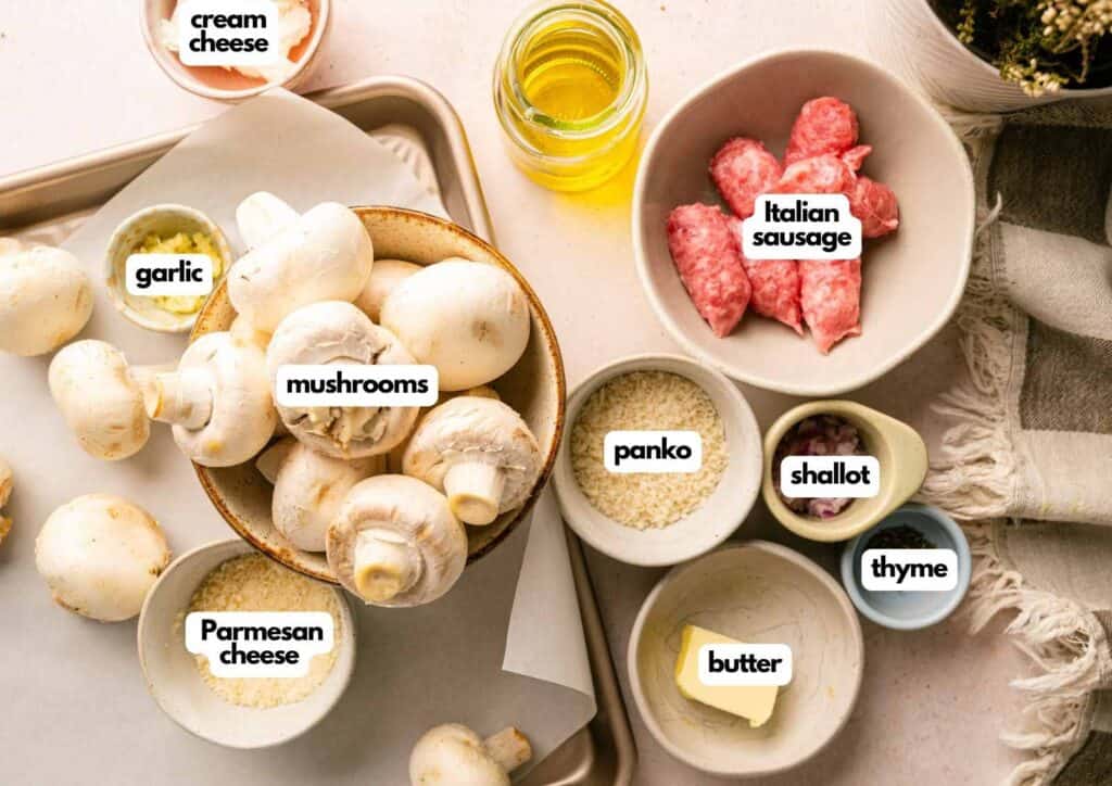 Ingredients for a recipe arranged in small bowls, including mushrooms, Italian sausage, cream cheese, panko, garlic, shallot, thyme, butter, and parmesan cheese.