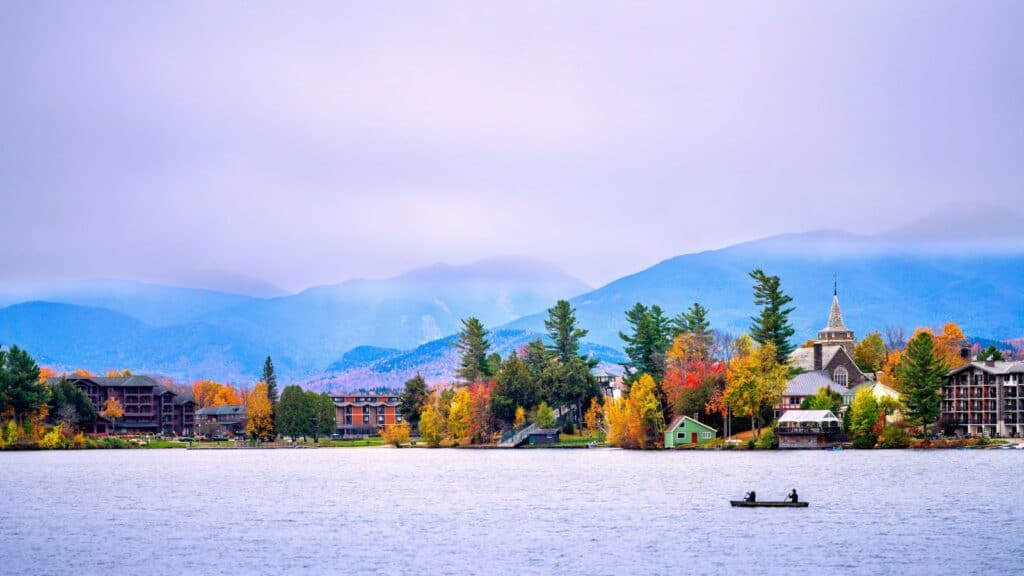 Two people in a canoe on a lake, framed by a village and autumn-colored trees perfect for fall-themed hotel packages in front of distant mountains.
