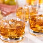 Two glasses of amber-colored whiskey with ice, sitting on a marble surface with crackers and grapes in the background.