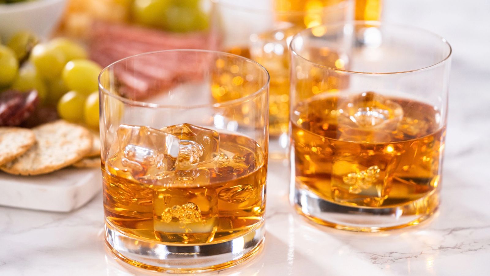 Two glasses of amber-colored whiskey with ice, sitting on a marble surface with crackers and grapes in the background.