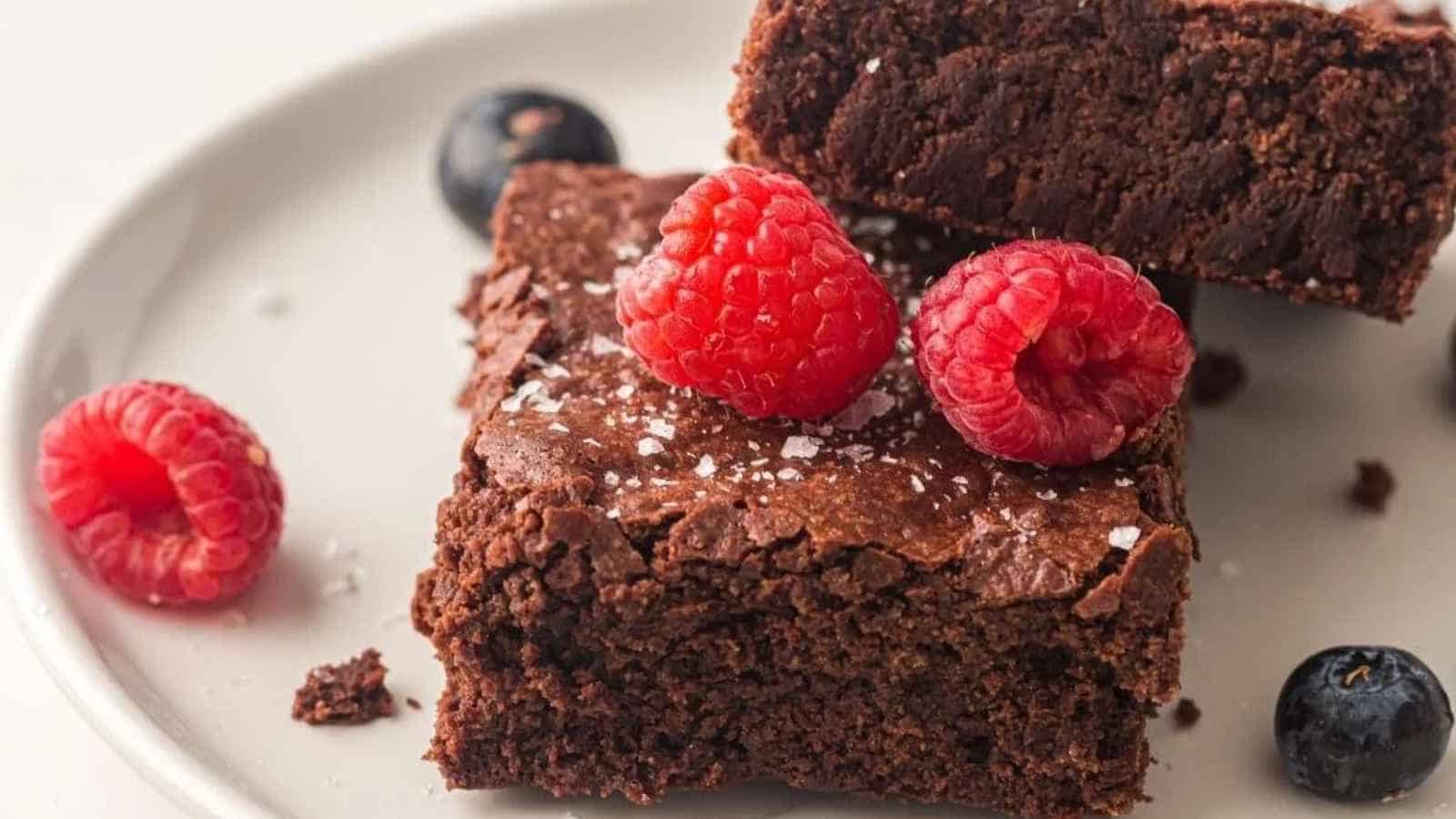 Two chocolate brownies topped with raspberries and coarse salt, served on a plate with fresh raspberries and blueberries.