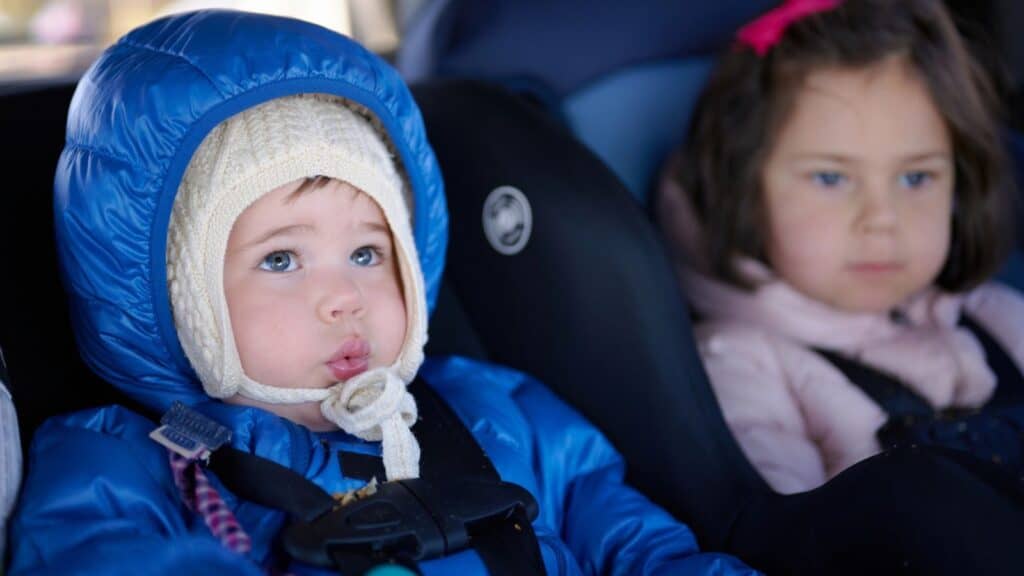 A baby in a blue hooded jacket and white cap sitting in a car seat next to a young girl, showcasing common fall closet-swap mistakes.