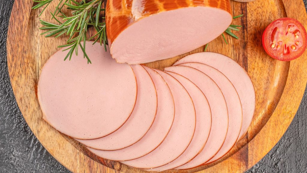 A sliced log of bologna on a wooden cutting board with rosemary and a tomato slice for National Bologna Day.
