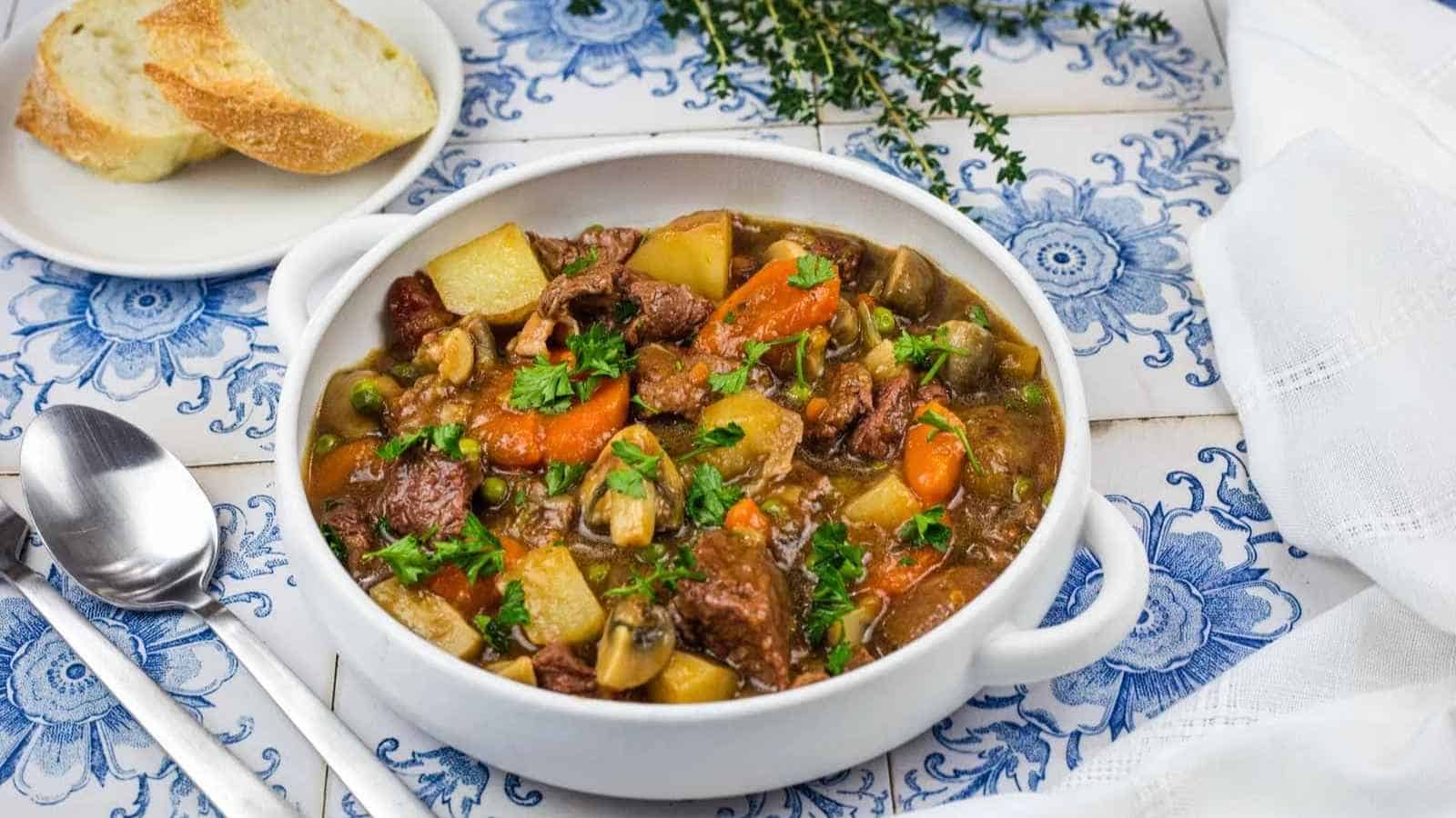 A bowl of beef stew with carrots, potatoes, and mushrooms, garnished with fresh herbs, served with sliced bread.