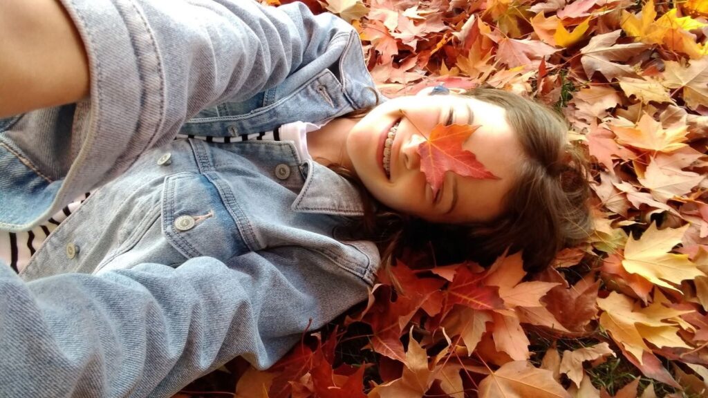A person in a denim jacket lies in a pile of autumn leaves, holding a maple leaf over one eye as part of their family fall traditions.
