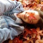 A person in a denim jacket lies in a pile of autumn leaves, holding a maple leaf over one eye as part of their family fall traditions.