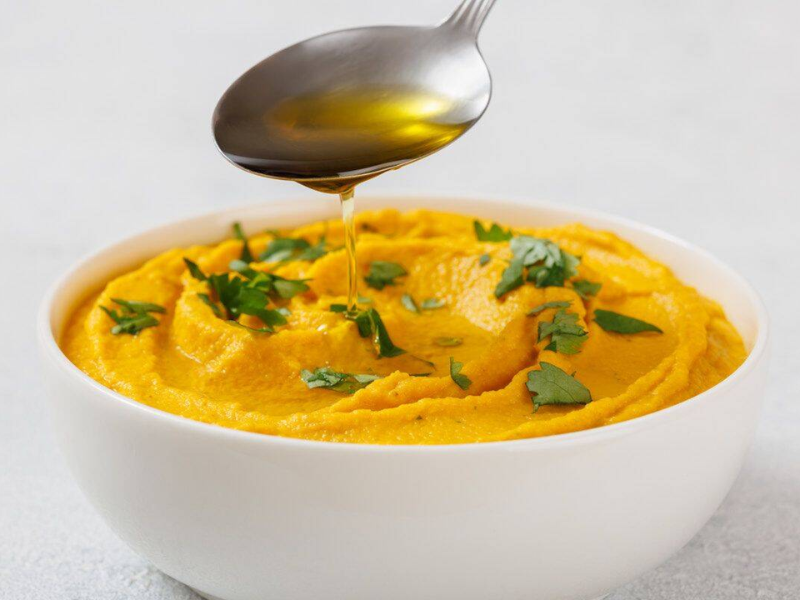 Bowl of Roasted Carrot Hummus garnished with green herbs, with a spoon drizzling oil above it.