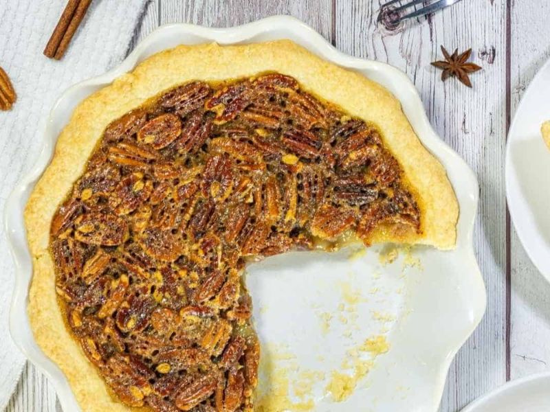 A pecan pie in a white dish with a large slice missing, shown on a light wooden surface.