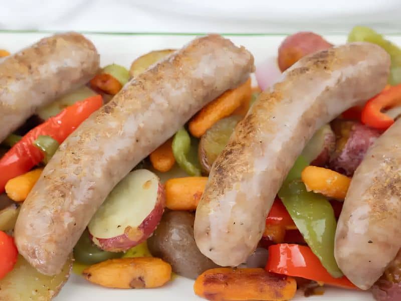 Four cooked sausages served on a bed of roasted vegetables, including carrots, red potatoes, green bell peppers, and red bell peppers.