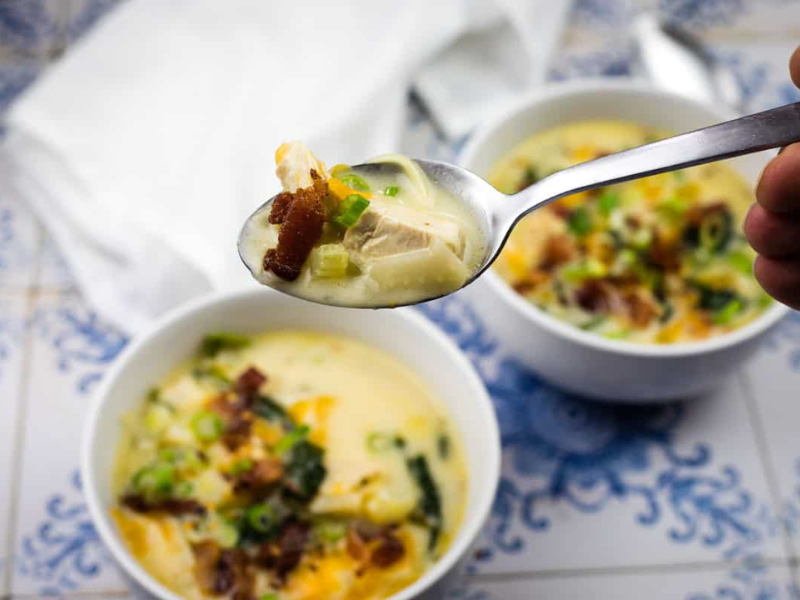 A hand holds a spoonful of Million dollar soup with chicken, bacon, and green onions above a bowl of soup on a blue and white tiled surface.
