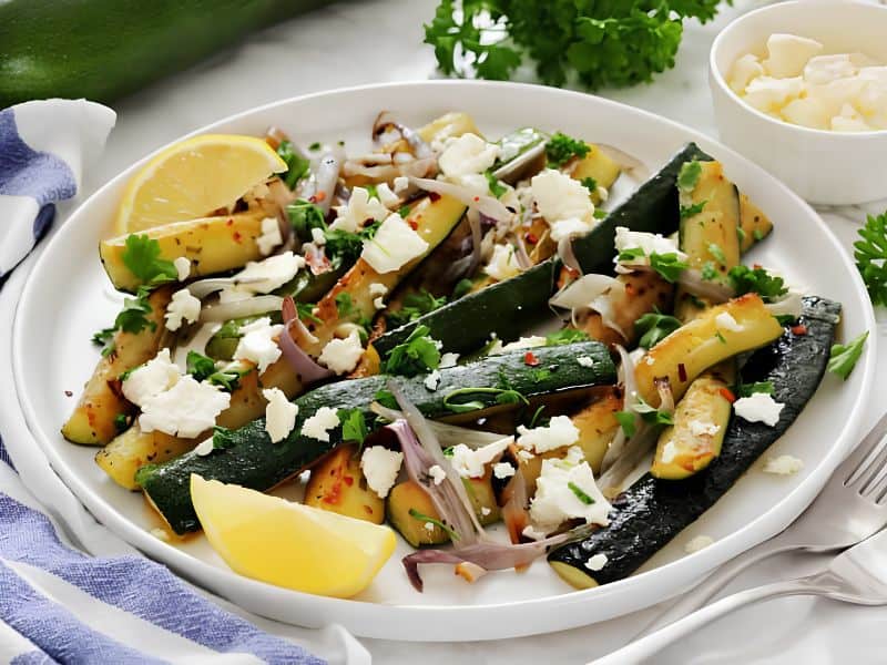 A plate of roasted zucchini and red onions garnished with crumbled cheese, herbs, red pepper flakes, and lemon wedges. A small bowl of cheese and fresh parsley are in the background.