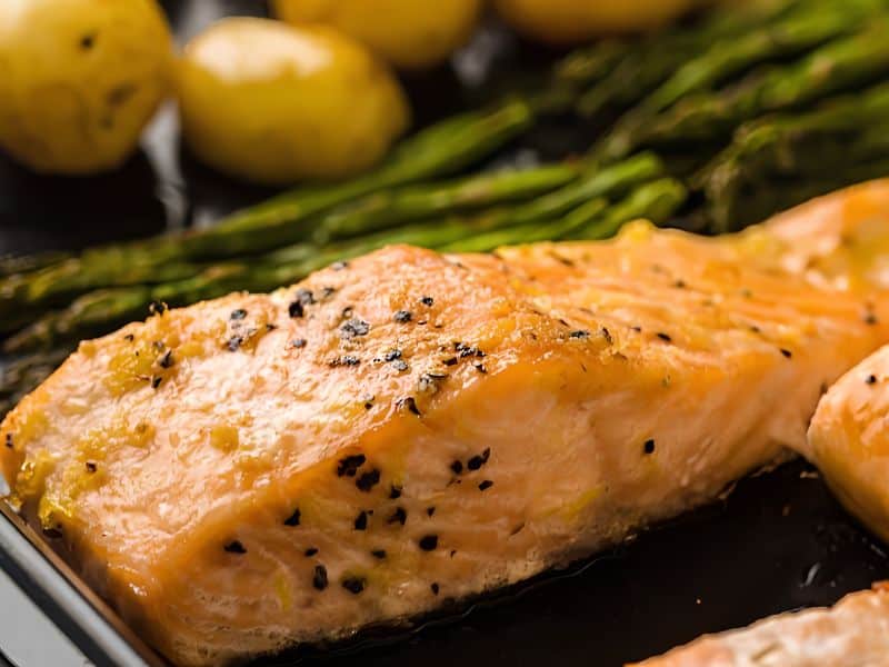Close-up of a baked salmon fillet seasoned with pepper, served alongside asparagus spears and small roasted potatoes.