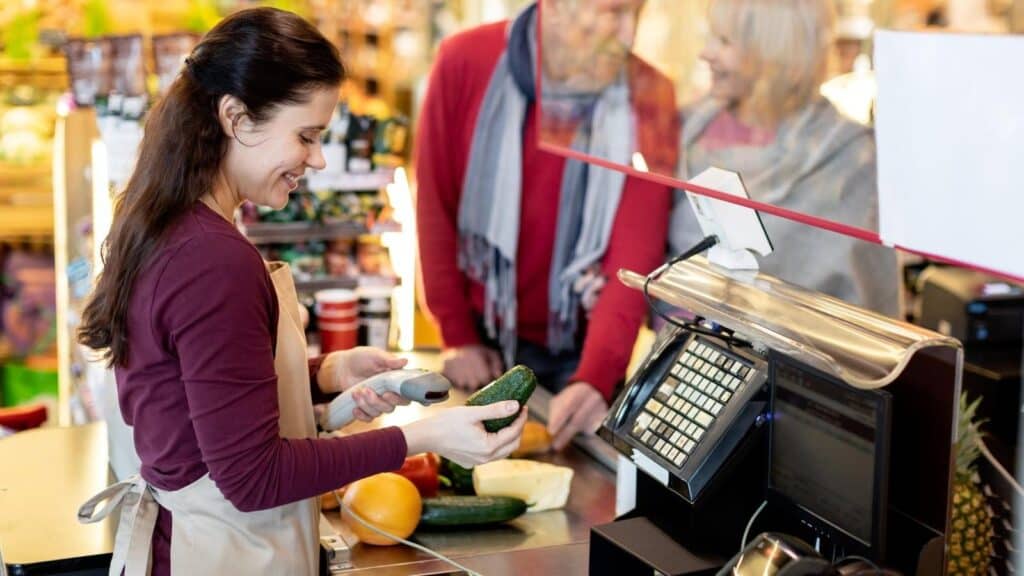 A cashier scans an avocado at a grocery store where Americans shop, while a customer waits at the checkout counter.