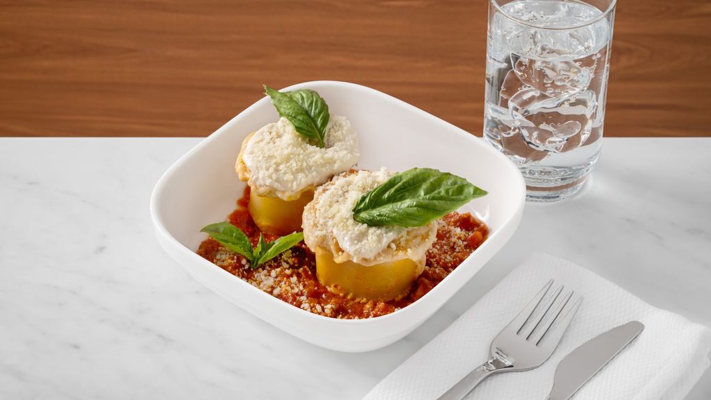 Two pasta roll-ups topped with cheese and basil in a bowl of tomato sauce, with a glass of water and silverware nearby.