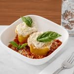 Two pasta roll-ups topped with cheese and basil in a bowl of tomato sauce, with a glass of water and silverware nearby.