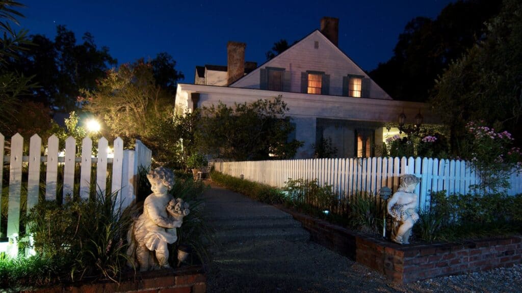 A haunted inn with illuminated windows seen at night beyond a white picket fence and two stone cherub statues flanking the walkway.