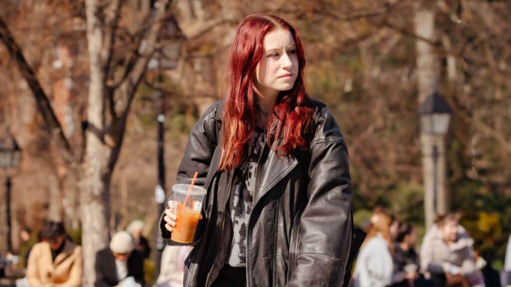 A person with long red hair wearing a black leather jacket holds an iced coffee in a public park, embracing the final days of the fall iced to hot coffee switch.
