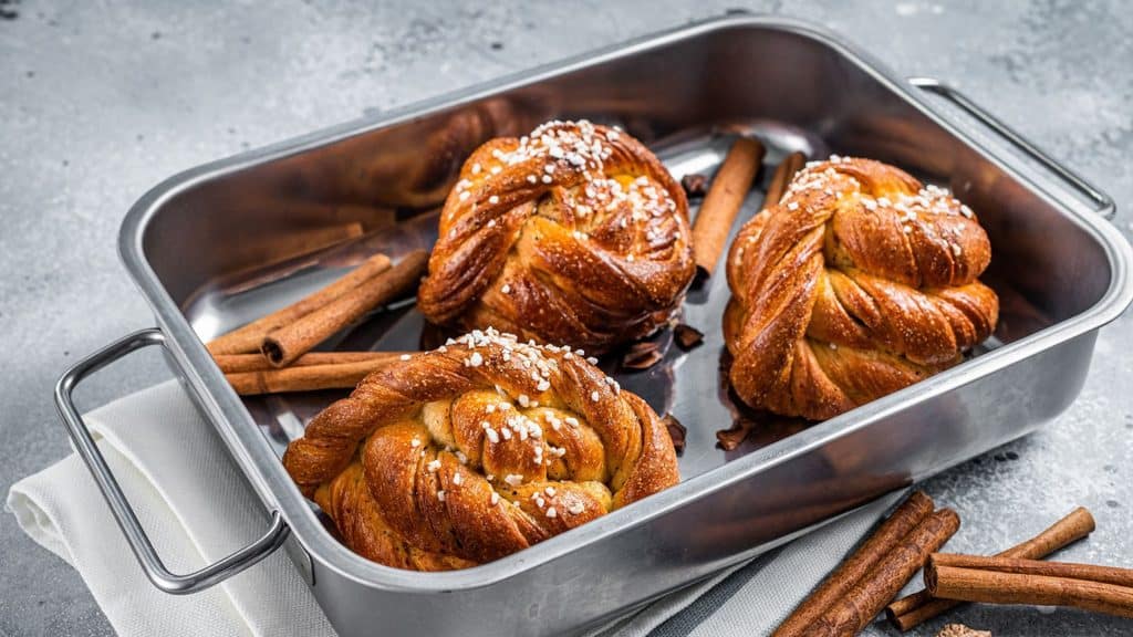Three braided cinnamon buns topped with pearl sugar in a metal baking pan, surrounded by cinnamon sticks.