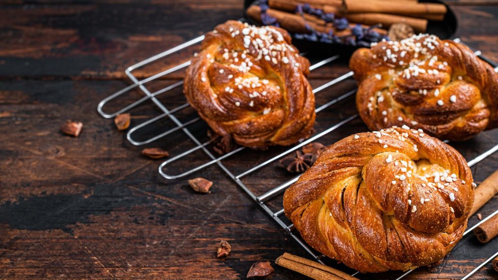 Three braided cinnamon buns topped with pearl sugar on a wire cooling rack.