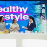 Three women stand at a counter of health products, similar to those found in QVC and HSN impulse buys, in front of a digital screen that reads “Healthy lifestyle.”.
