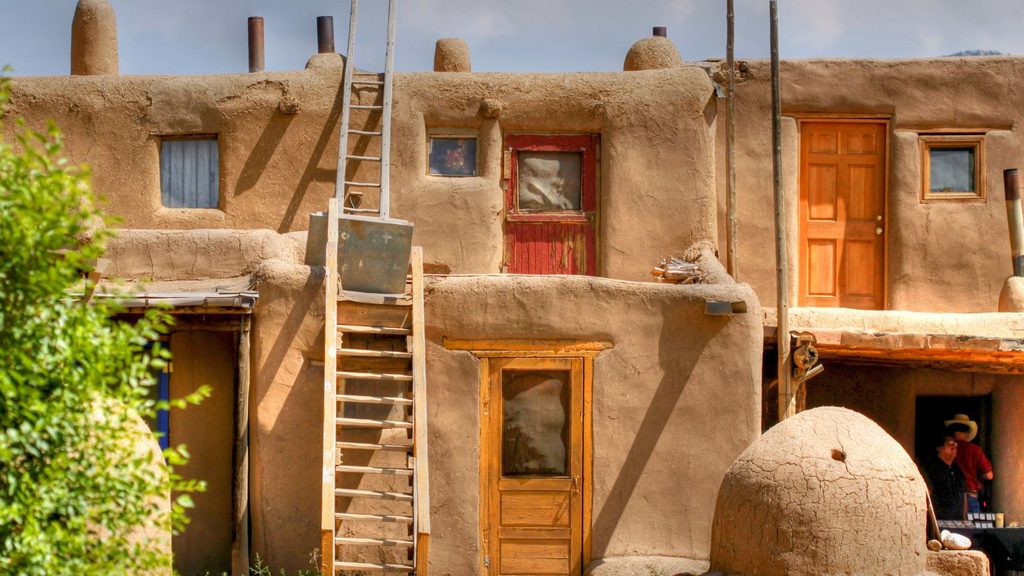 Two-story adobe building with wooden ladders, doors, and a beehive-shaped oven in the foreground, evoking the history of Taos food.