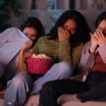 Three people sitting on a couch, looking frightened while watching scary movies for a marathon with a bowl of popcorn.