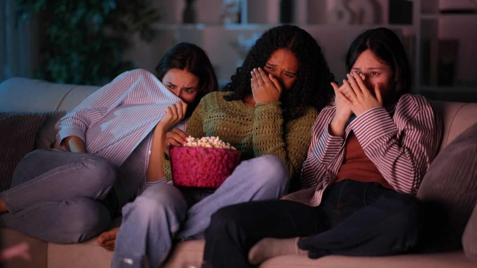 Three people sitting on a couch, looking frightened while watching scary movies for a marathon with a bowl of popcorn.