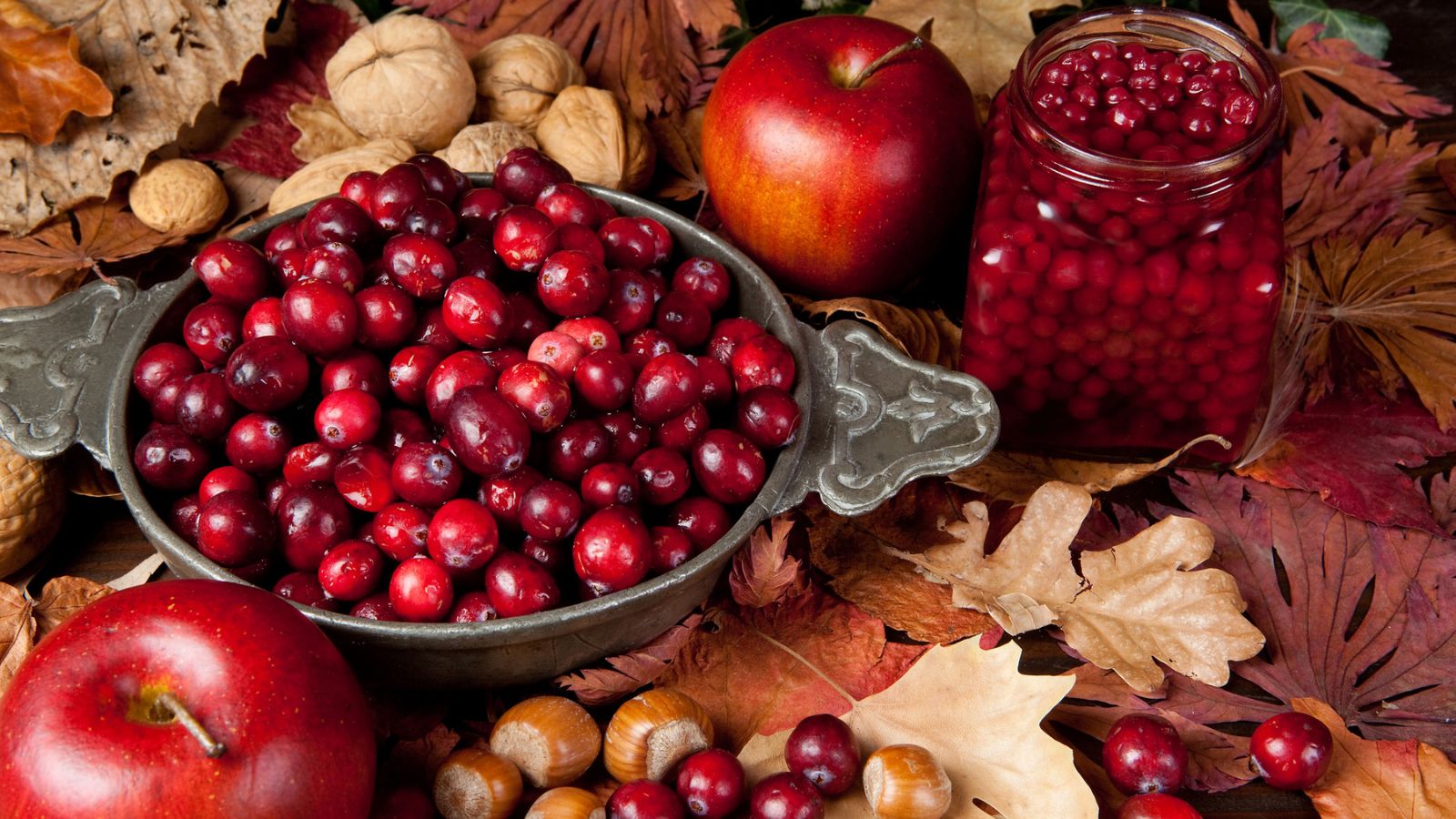 A metal bowl of fresh cranberries, a jar of cranberry sauce, red apples, and assorted nuts arranged on a bed of dried autumn leaves as part of a collection of Thanksgiving groceries.