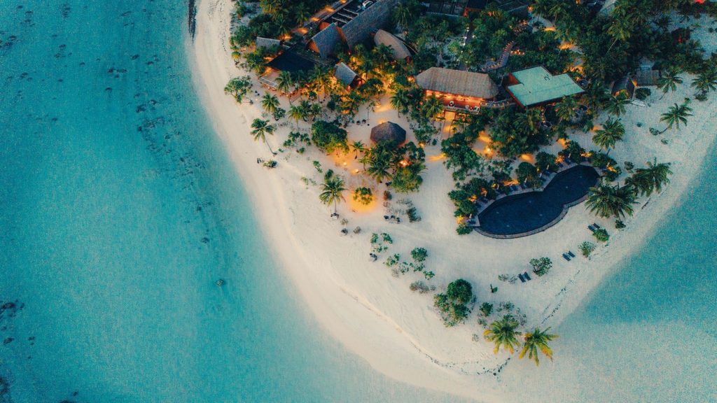 Aerial view of a tropical island resort with thatched-roof buildings and a pool illuminated at twilight, bordered by a sandy beach and turquoise water.