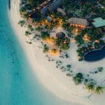 Aerial view of a tropical island resort with thatched-roof buildings and a pool illuminated at twilight, bordered by a sandy beach and turquoise water.