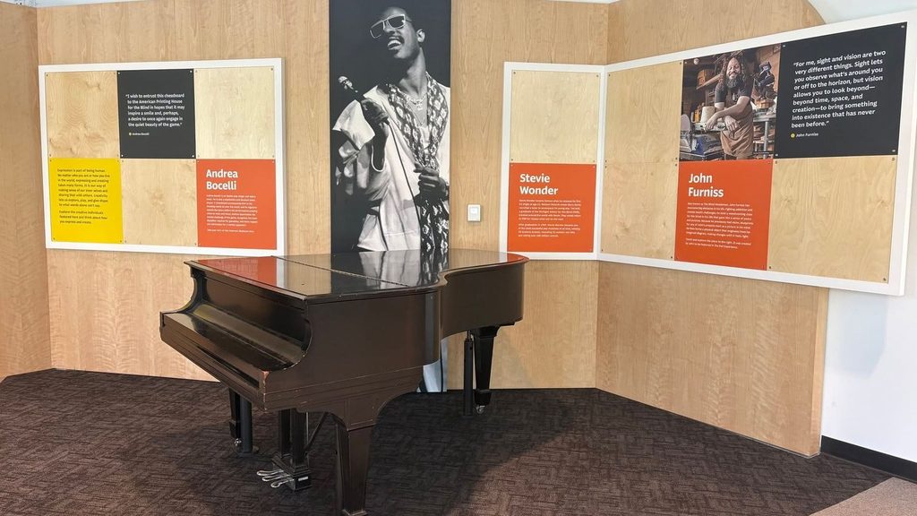 A grand piano sits in a Louisville museum display featuring photographs and biographies of Andrea Bocelli, Stevie Wonder, and John Furniss.
