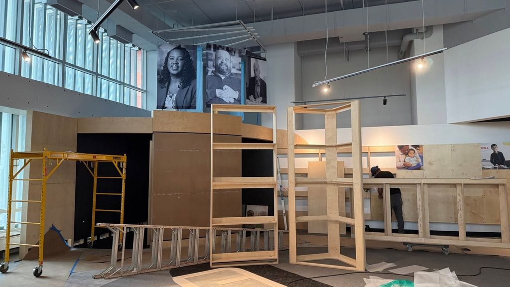 A construction site in a Louisville museum exhibition space featuring plywood structures, a yellow scaffolding tower, ladders, and portrait posters on the back wall.