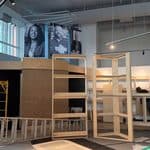A construction site in a Louisville museum exhibition space featuring plywood structures, a yellow scaffolding tower, ladders, and portrait posters on the back wall.