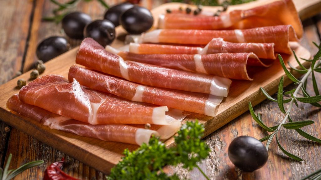 Thinly sliced prosciutto arranged on a wooden serving board with black olives and fresh herbs.