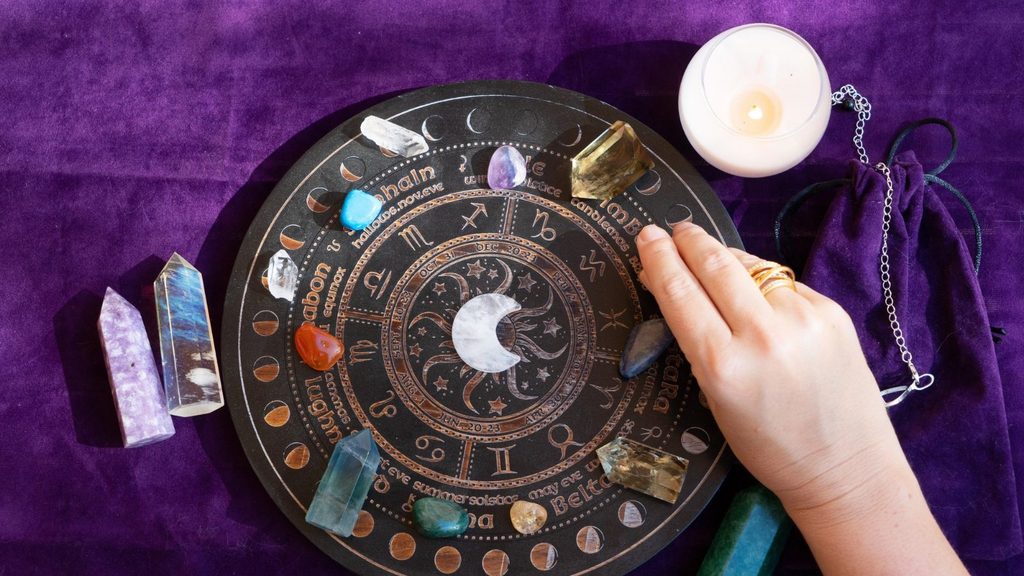 A hand rests on a circular astrology calendar board decorated with crystals, a lit candle, and a velvet bag on a purple cloth during an astrology wellness retreat.