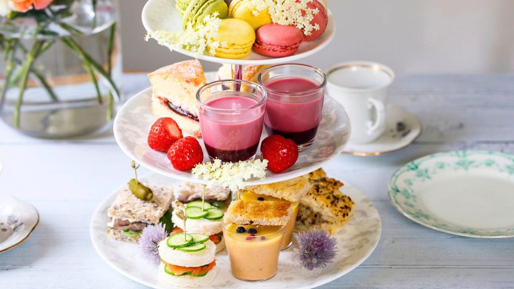 A three-tiered afternoon tea stand for National Dessert Day featuring macarons, cake, berry mousse, fresh strawberries, finger sandwiches, and small cups of soup.
