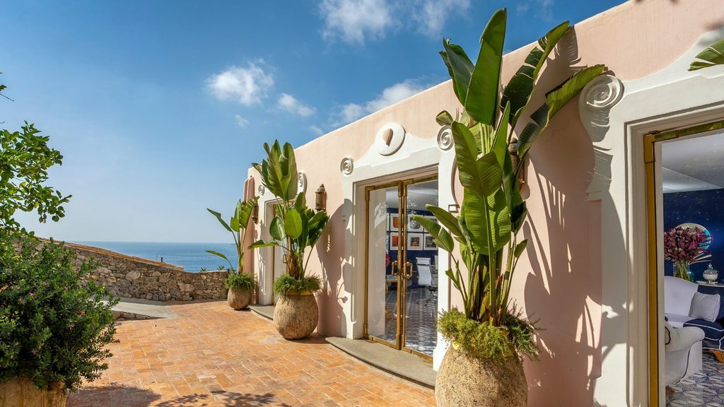 A pink exterior wall with glass doors and potted bird of paradise plants on a brick terrace overlooking the ocean, reflecting luxury hotel openings 2027.