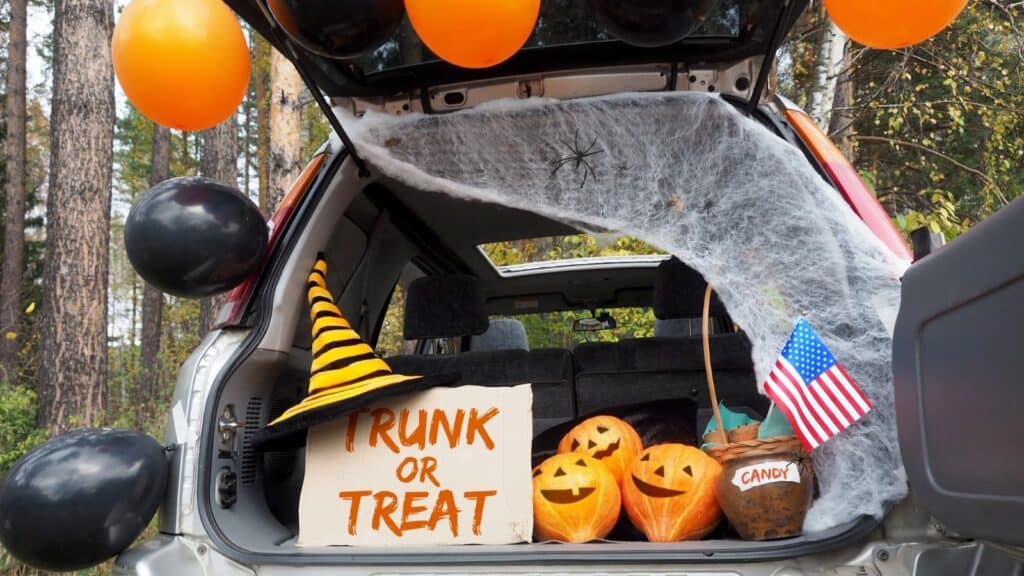 An open car trunk decorated for a trunk-or-treat event with balloons, fake spiderwebs, carved pumpkins, a witch hat, and a small American flag to display trunk-or-treat snack ideas.