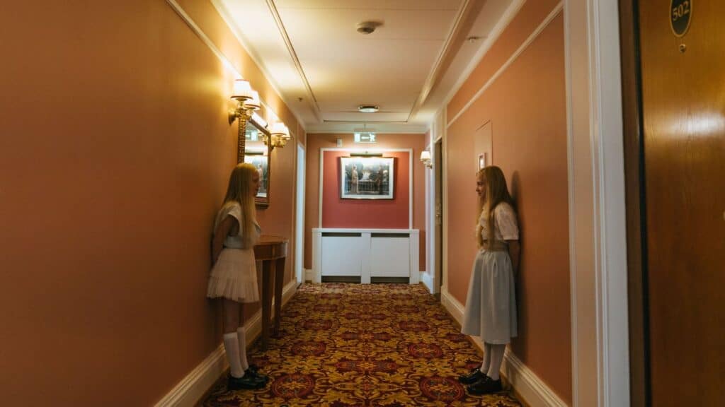 Two young girls in matching white dresses and knee-high socks stand facing each other in a patterned hotel hallway, capturing the vibe of a spookiest small-town fall road trip.