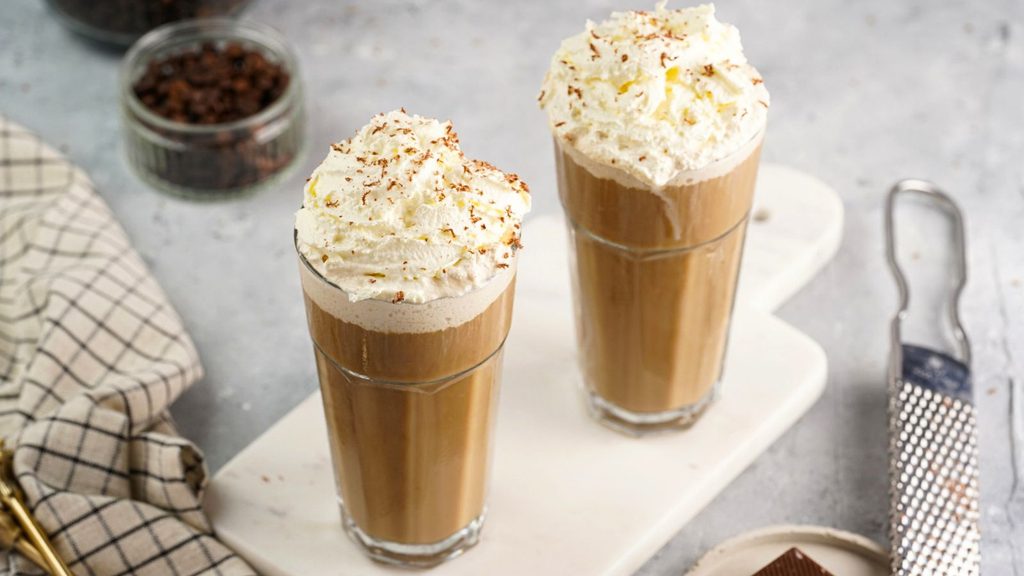 Two tall glasses of iced coffee, perfect for National Frappe Day, topped with whipped cream and chocolate shavings on a white serving board.