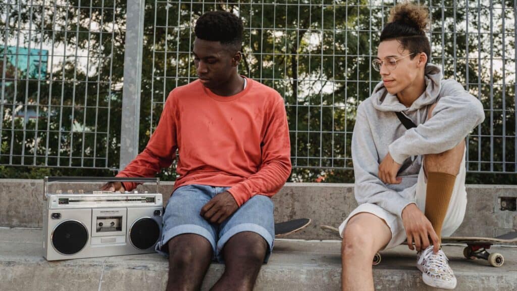 Two young men sit on a concrete ledge with skateboards and a boombox, evoking boom box memories.