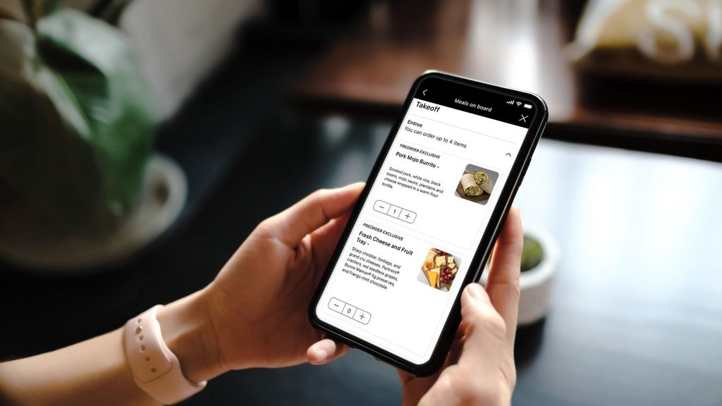 A person holding a smartphone displaying an inflight meal ordering app with options for a pork burrito and a cheese and fruit tray.