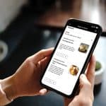 A person holding a smartphone displaying an inflight meal ordering app with options for a pork burrito and a cheese and fruit tray.